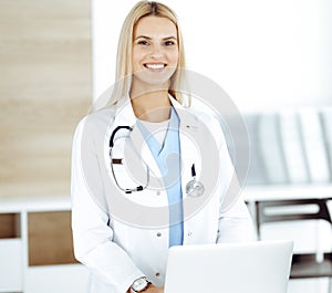 Blond female physician is lsmiling while using laptop computer. Woman-doctor at work in clinic excited and happy of her