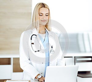 Blond female physician is lsmiling while using laptop computer. Woman-doctor at work in clinic excited and happy of her