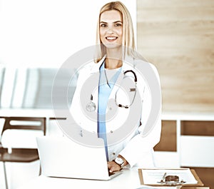 Blond female physician is lsmiling while using laptop computer. Woman-doctor at work in clinic excited and happy of her