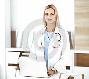 Blond female physician is lsmiling while using laptop computer. Woman-doctor at work in clinic excited and happy of her