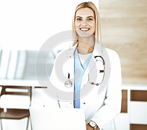 Blond female physician is lsmiling while using laptop computer. Woman-doctor at work in clinic excited and happy of her