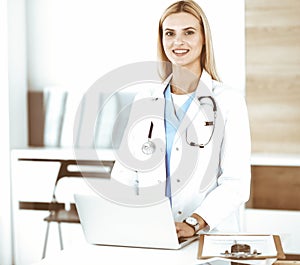 Blond female physician is lsmiling while using laptop computer. Woman-doctor at work in clinic excited and happy of her