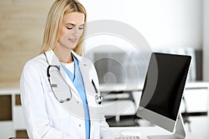 Blond female physician is standing at her workplace near desktop computer. Woman-doctor is excited and happy of her