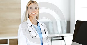 Blond female physician is standing at her workplace near desktop computer. Woman-doctor is excited and happy of her