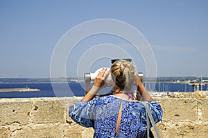 Blond female tourist looks through binoculars at the sea from Gallipoli castle. Apulia, Italy