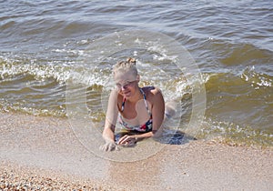 Blond girl in a bikini lying on the beach and the waves splash on it. Beautiful young woman in a colorful bikini on sea background