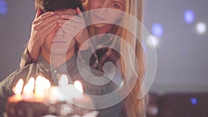 Blond girl covering eyes of the man, then let him see the cake with many candles. The guy has birthday celebration