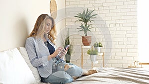 Blond girl in jeans sits on bed working on smartphone