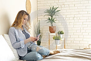 Blond girl in jeans sits on bed working on smartphone