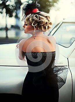 Blond girl model with bright makeup and curly hairstyle in retro style posing near old white car