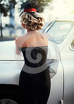 Blond girl model with bright makeup and curly hairstyle in retro style posing near old white car