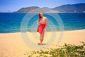 blond girl in red stands barefoot on sand beach at noon