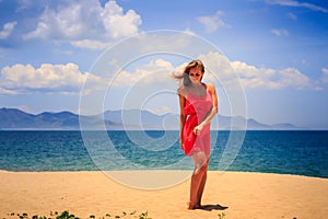 blond girl in red stands on sand holds hand on hip at noon