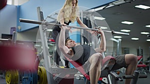 Blond gorgeous female personal trainer endorsing masculine handsome sportsman lifting barbell. Beautiful woman