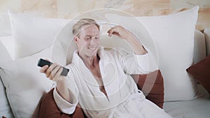 Blond guy in bathrobe switches on TV on comfortable bed