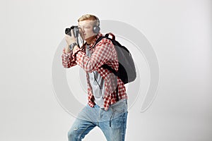 Blond guy in headphones, with black backpack on his shoulder dressed in a white t-shirt, red checkered shirt and jeans