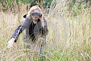 Blond hair European woman spying with binoculars, move to the side high dry grass, black jacket