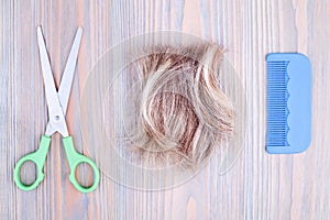 Blond hair lock, metal scissors, plastic comb light wooden background close up, cut off blonde hair curl on bright wood, shears