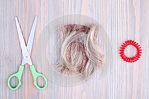 Blond hair lock, scissors, scrunchie light wooden background close up, cut off blonde hair curl on bright wood, shears, hair band