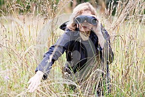 Blond hair mature woman spying with binoculars, move to the side dry grass, black clothing