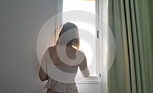 Blond hair young woman standing in the sunrise with harmony inside the room, looking through the window in casual clothes. Sexy