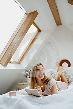 Blond happy woman making notes in note pad while sitting on bed at home