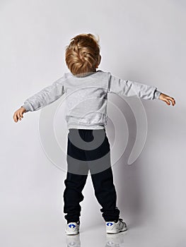 Blond kid boy in black pants and sweatshirt stands back to camera holding head up and arms spread wide