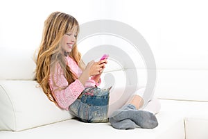 Blond kid girl playing fun with mobile phone on white sofa