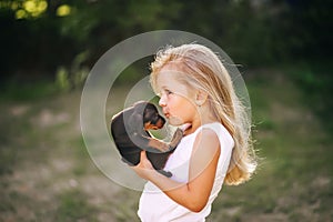 Blond kid girl in white dress is kissing puppy pet dog outdoors. Side view