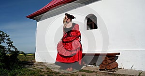 Blond lady in red costume dress sitting in nature near chapel relaxing