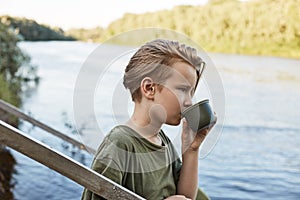 Blond little boy drinking hot coffee or tea from thermos isolated over nature landscape, male child spending time in open air, guy