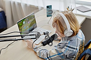 Blond little boy in headphones speaking in microphone while sitting by desk