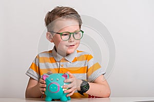 Blond little boy putting money on a moneybox. Portrait of a stylish kid in green glasses with watch and piggy bank in