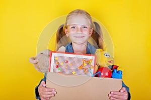 Blond little girl holding a box with toys on yellow background. Donation concept..