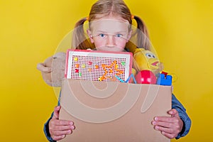 Blond little girl holding a box with toys on yellow background. Donation concept..