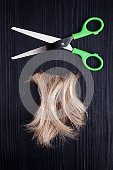 Blond lock of hair, metal open scissors black wooden background close up, cut off natural blonde hair curl on dark wood, shears