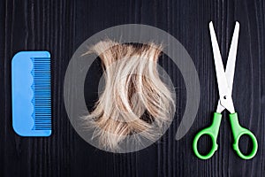 Blond lock of hair, metal scissors, plastic comb black wooden background closeup, cut off blonde hair curl on dark wood, shears