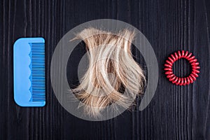 Blond lock of hair, red scrunchie, blue plastic comb black wooden background closeup, cut off blonde hair curl on dark wood, hair
