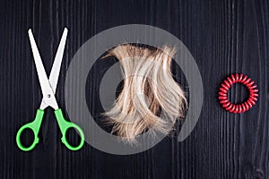 Blond lock of hair, scissors, scrunchy black wooden background closeup, cut off blonde hair curl on dark wood, shears, spiral band