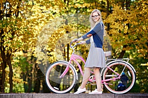 Blond long-haired attractive girl on pink lady bicycle in sunny autumn park on trees background.