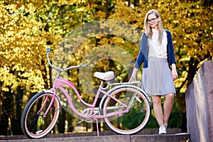 Blond long-haired attractive girl on pink lady bicycle in sunny autumn park on trees background.