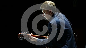 Blond man playing acoustic guitar on stage at spotlight, unplugged performance. back view