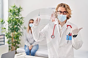 Blond man wearing doctor uniform and medical mask holding syringe pointing thumb up to the side smiling happy with open mouth