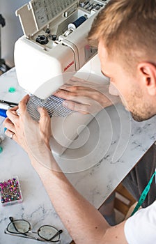 A blond man in white T-shirt is sewing in home workshop. A seamster with a measuring tape on his neck. The concept of small