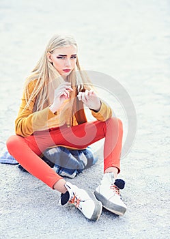 Blond model with thick eyebrows and blushed cheeks sitting on sandy road. Girl in bright outfit posing outdoors. Street