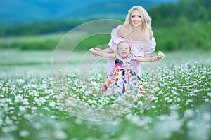 Blond mother with cute little daughter wearing white colourful pink dresses in chamomile field, summer time Enjoying spending time