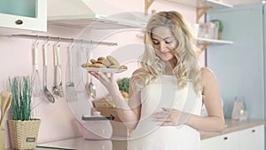 Blond pregnant woman holding plate with healthful sweet cookies and smiling at camera. Portrait of young beautiful