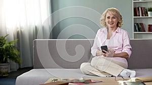 Blond senior woman smiling in camera, holding smartphone, app online service