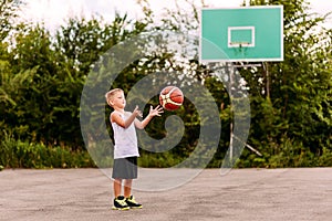A blond seven-year-old boy in a basketball uniform throws a ball on an open basketball court in the summer. Children and sports