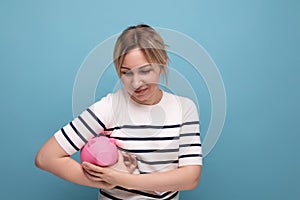 blond smiling girl in casual outfit holding a piggy bank on a blue background with copy space